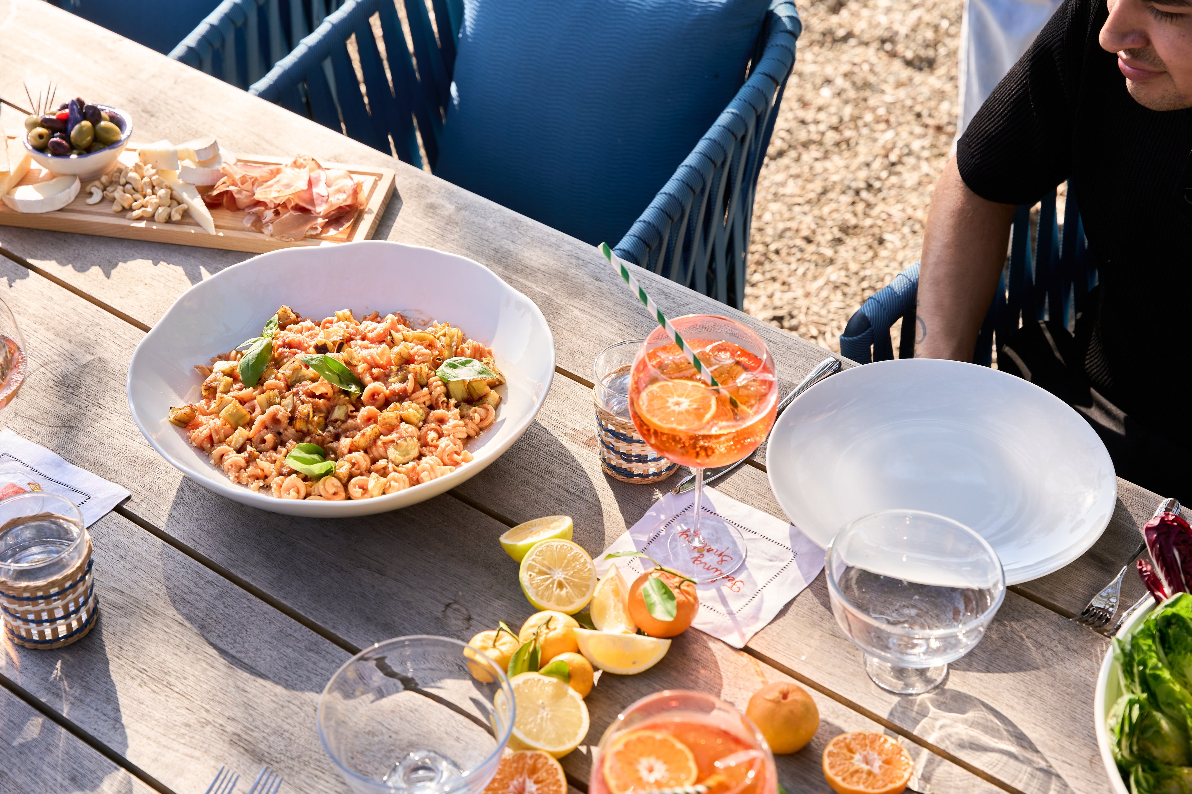 Outdoor setting with a table full of pasta and other food, drinks, and a person sitting nearby.