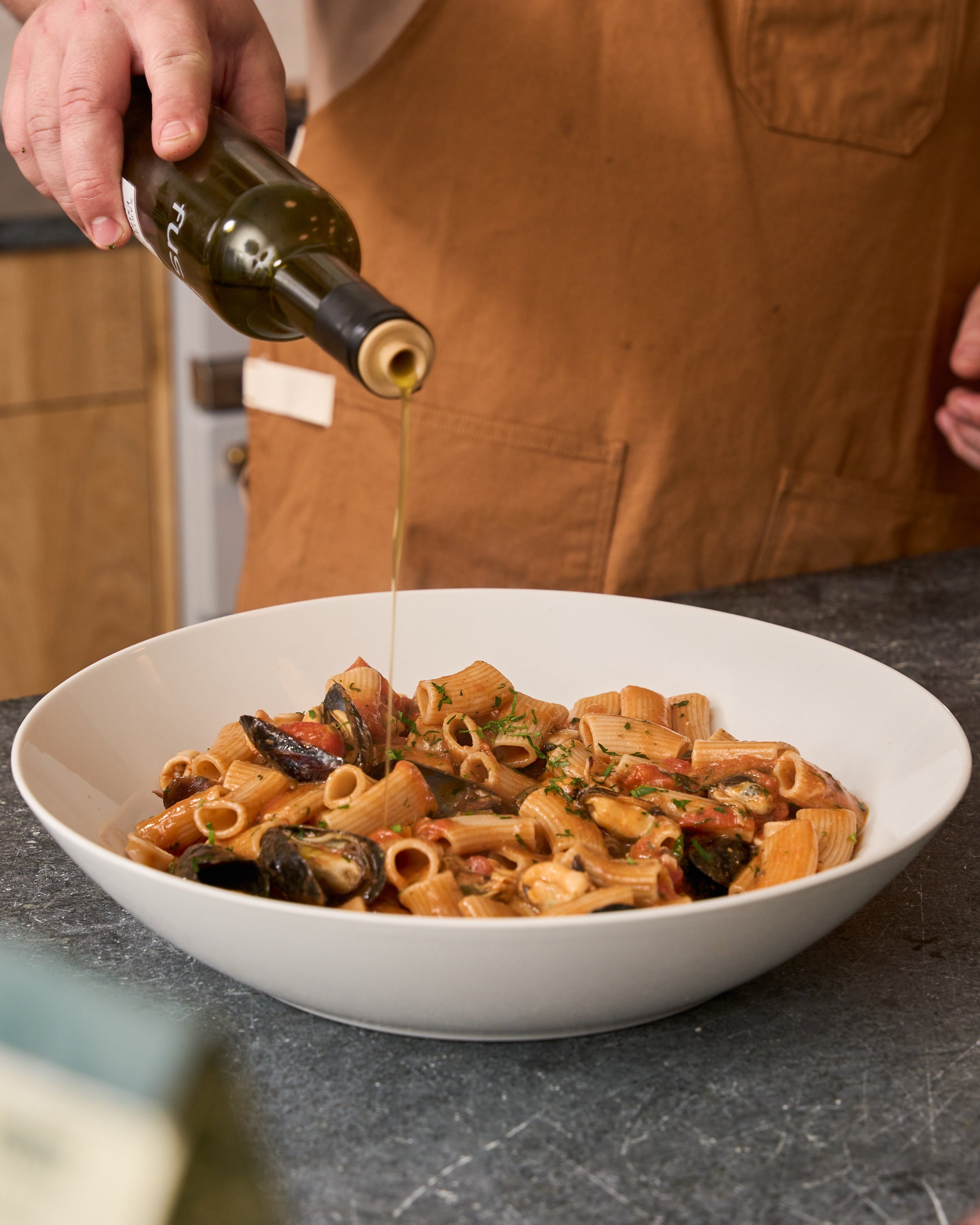 Person pouring olive oil over a bowl of pasta on a kitchen counter